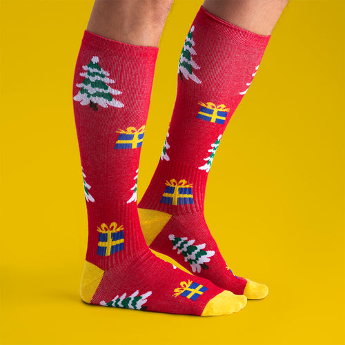 Röda stödstrumpor från Svenskhusman med mönster av julgranar och paket mot en gul bakgrund.