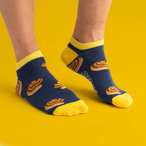 Blå och gula sneakersstrumpor mönstrade med kanelbullar från Svenskhusman på fötter mot en gul bakgrund.