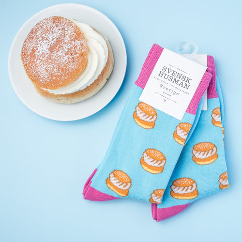 Ljusblå strumpor med semlemotiv och rosa detaljer bredvid en klassisk semla - Svenskhusman