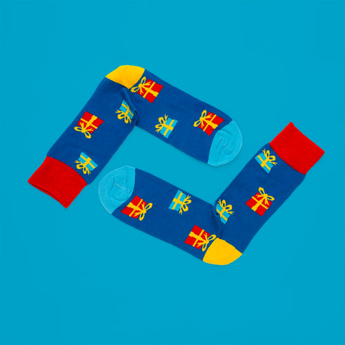 Blå julstrumpor med mönster av färgglada paket, röda muddar och gula detaljer från Svenskhusman.