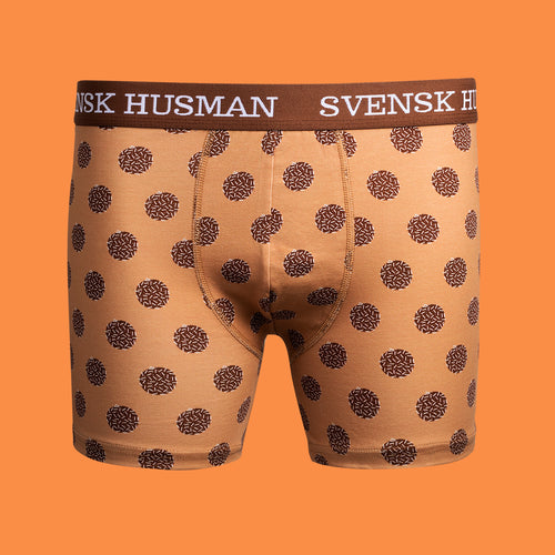 Ljusbruna boxerkalsonger med chokladbollsmönster och logotypresår från Svenskhusman på orange bakgrund.