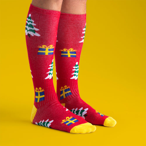 Röda stödstrumpor med mönster av julgranar och paket från Svenskhusman mot en gul bakgrund.