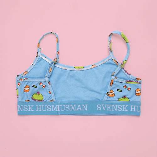 Ljusblå BH-topp med fika-mönster av bakelser och resårband med Svensk Husman-logotyp mot rosa bakgrund.