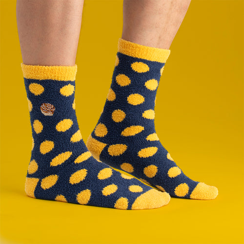 Fluffy blue socks with yellow polka dots and an embroidered cinnamon bun on feet against a yellow background.
