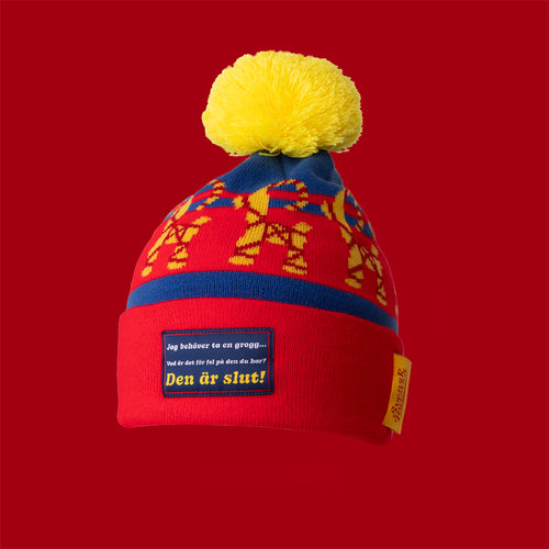 Röd och blå stickad mössa med gul tofs och texten Jag behöver ta en grogg, från Svenskhusman.