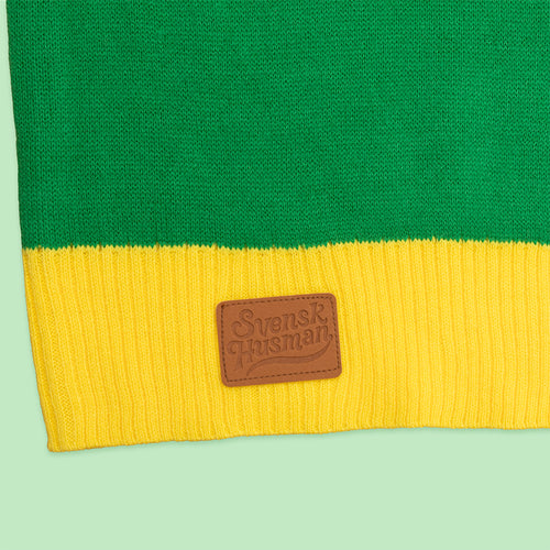 Närbild på grön och gul stickad jultröja med Svenskhusman-logotyp på en brun läderlapp.