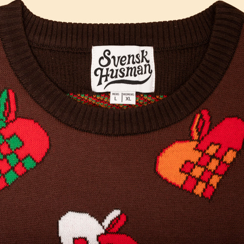 Brun stickad jultröja med mönster av julhjärtan och vit Svenskhusman-etikett i halsringningen.
