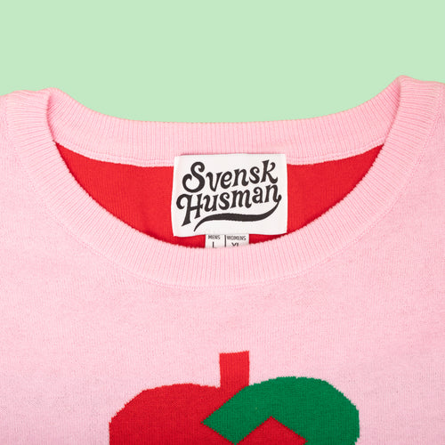 Rosa stickad jultröja med ett rött julhjärta och vit etikett från Svenskhusman på grön bakgrund.