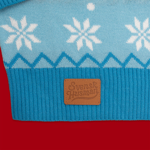 Blau stickad jultröja med weißa snöflingor och en braun Schwedischhusman-logotyp mot rot bakgrund.