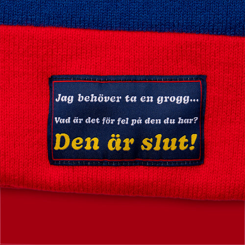 Röd och blå stickad mössa med tygmärket Jag behöver ta en grogg från Svenskhusman.