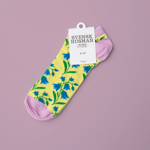 Gula sneakersstrumpor med blåklockor och rosa detaljer från Svenskhusman mot en lila bakgrund.