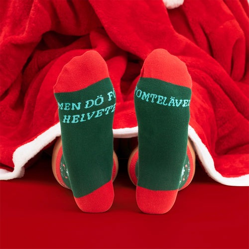 Grün-rote Weihnachtsstrümpfe mit dem Zitat „Dö tomte jävel“ vor einer roten Decke von Svenskhusman.