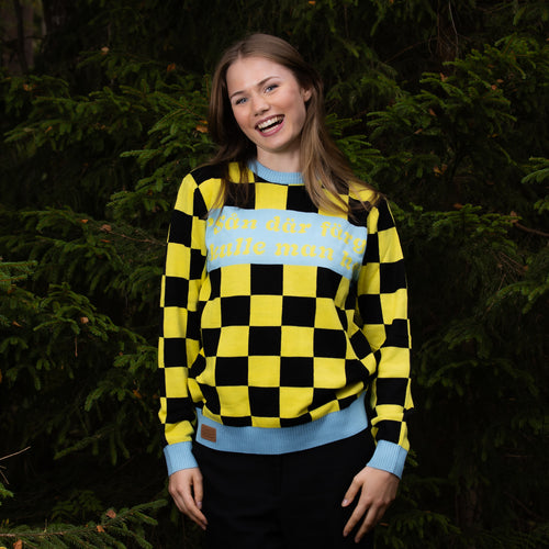 Yellow checkered Svenskhusman Christmas sweater with Swedish text worn by a woman in a forest.