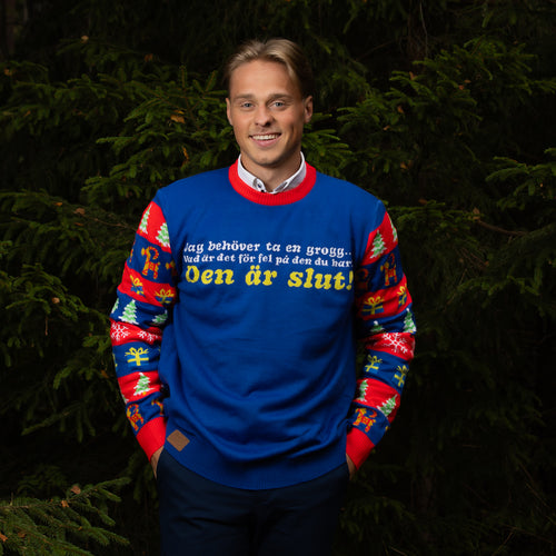 Blue Svenskhusman Christmas sweater with humorous Swedish text worn by a man in a spruce forest.