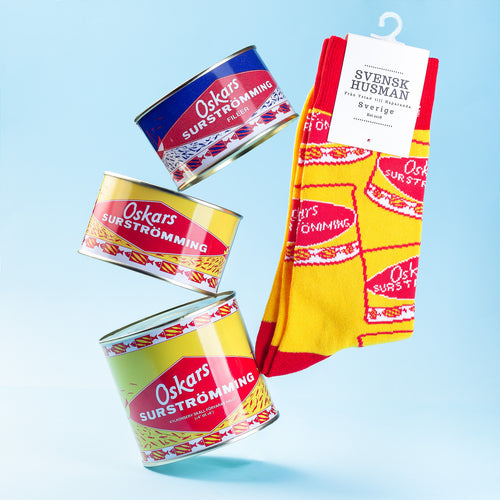 Gula och röda strumpor med mönster av Oskars Surströmming intill matchande konservburkar - Svenskhusman