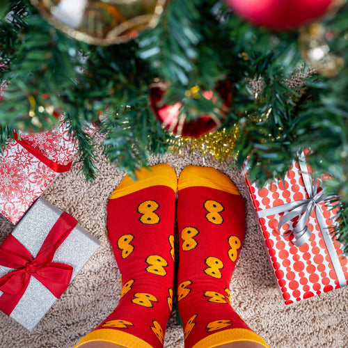 Red Christmas socks with a yellow saffron bun pattern on feet under a festive tree with wrapped presents.