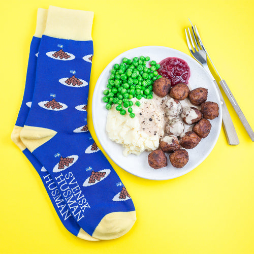 Blå och gula strumpor med köttbullsmotiv intill en tallrik med klassisk husmanskost på gul bakgrund.