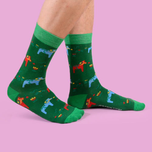 Green socks with colorful Dala horses worn by a person against a pink background - Svenskhusman.