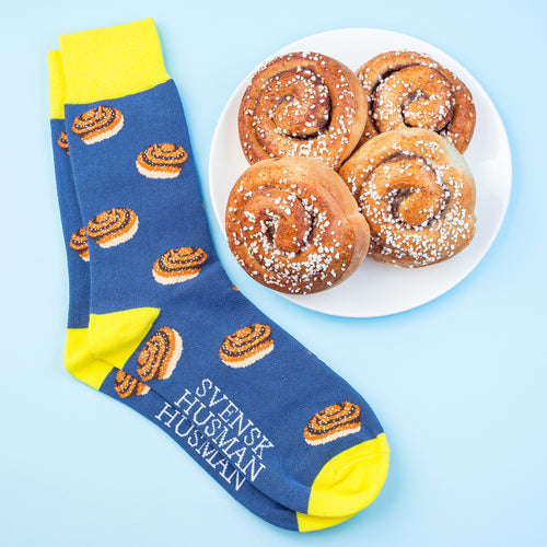 Blå och gula mönstrade strumpor med kanelbullar bredvid en tallrik med bullar från Svenskhusman.
