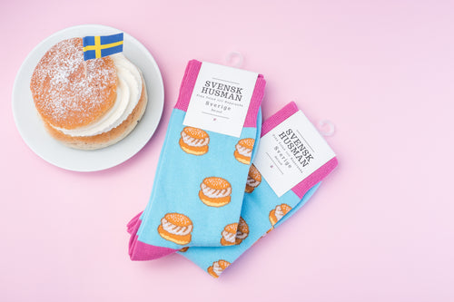 Turquoise socks with semla motifs next to a semla pastry with a Swedish flag on a pink background.