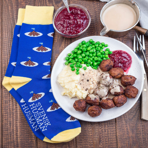 Blå och gula strumpor med köttbullsmönster från Svenskhusman bredvid en tallrik med klassisk husmanskost.